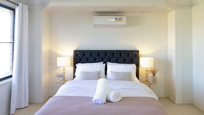 A hotel room with a large bed, two bedside lamps, a wall-mounted air conditioner, and a window with curtains. - Spacious, Sunlit & Recently Renovated Family Home - 4BD & Hotel Quality Bedding (Prestons)