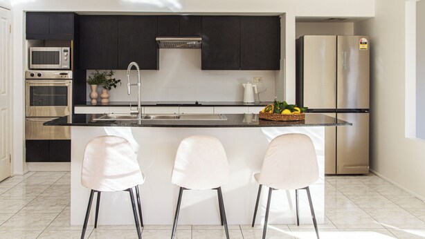 A modern kitchen with dark cabinets, a stainless steel refrigerator, a microwave, and a sink. There are three white bar stools and a small island with a fruit bowl. - Spacious, Sunlit & Recently Renovated Family Home - 4BD & Hotel Quality Bedding (Prestons)