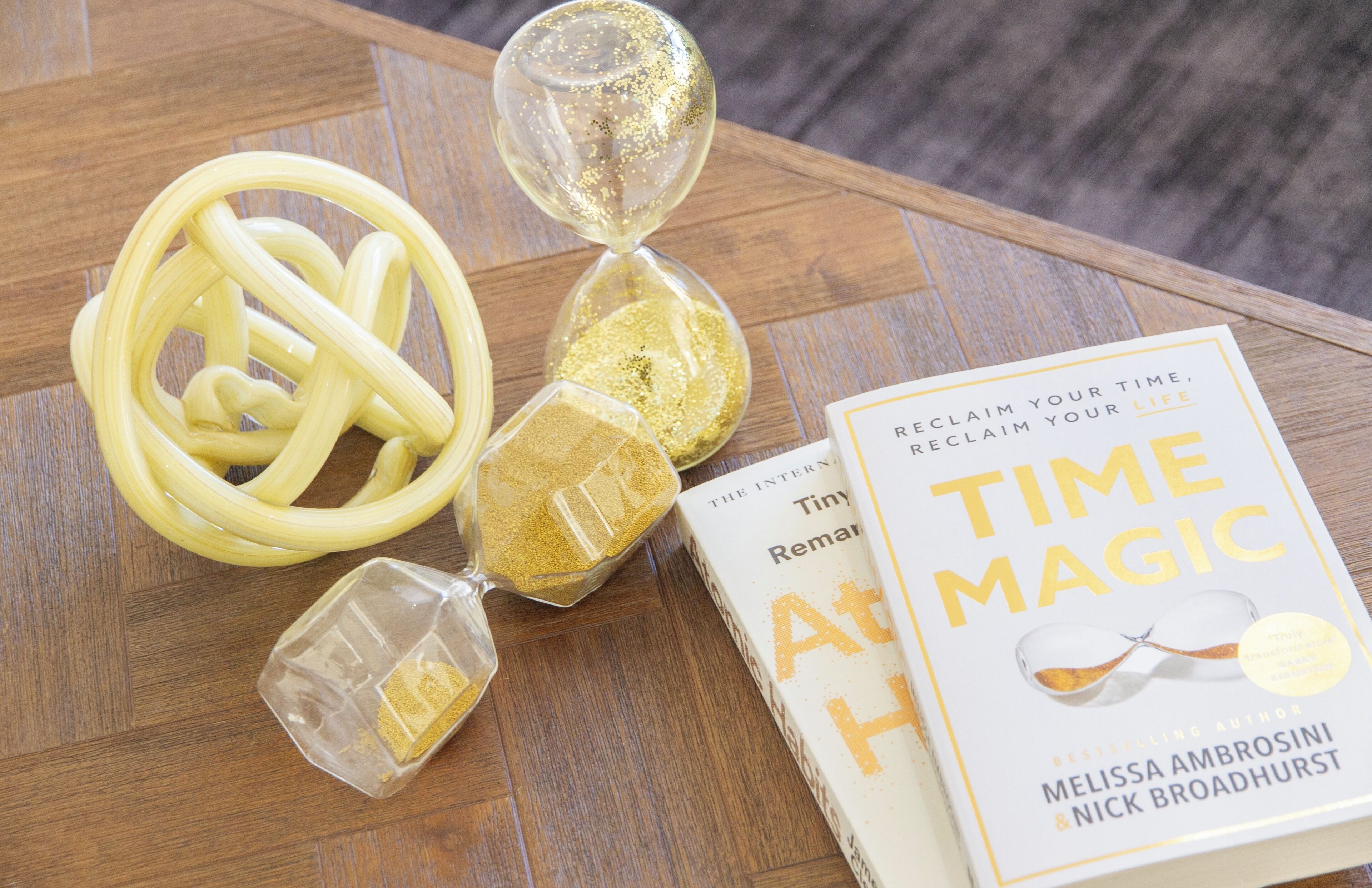 A book titled "Time Magic" by Melissa Ambrosini and Nick Broadhurst, a golden hourglass, and a yellow coiled object on a wooden surface.