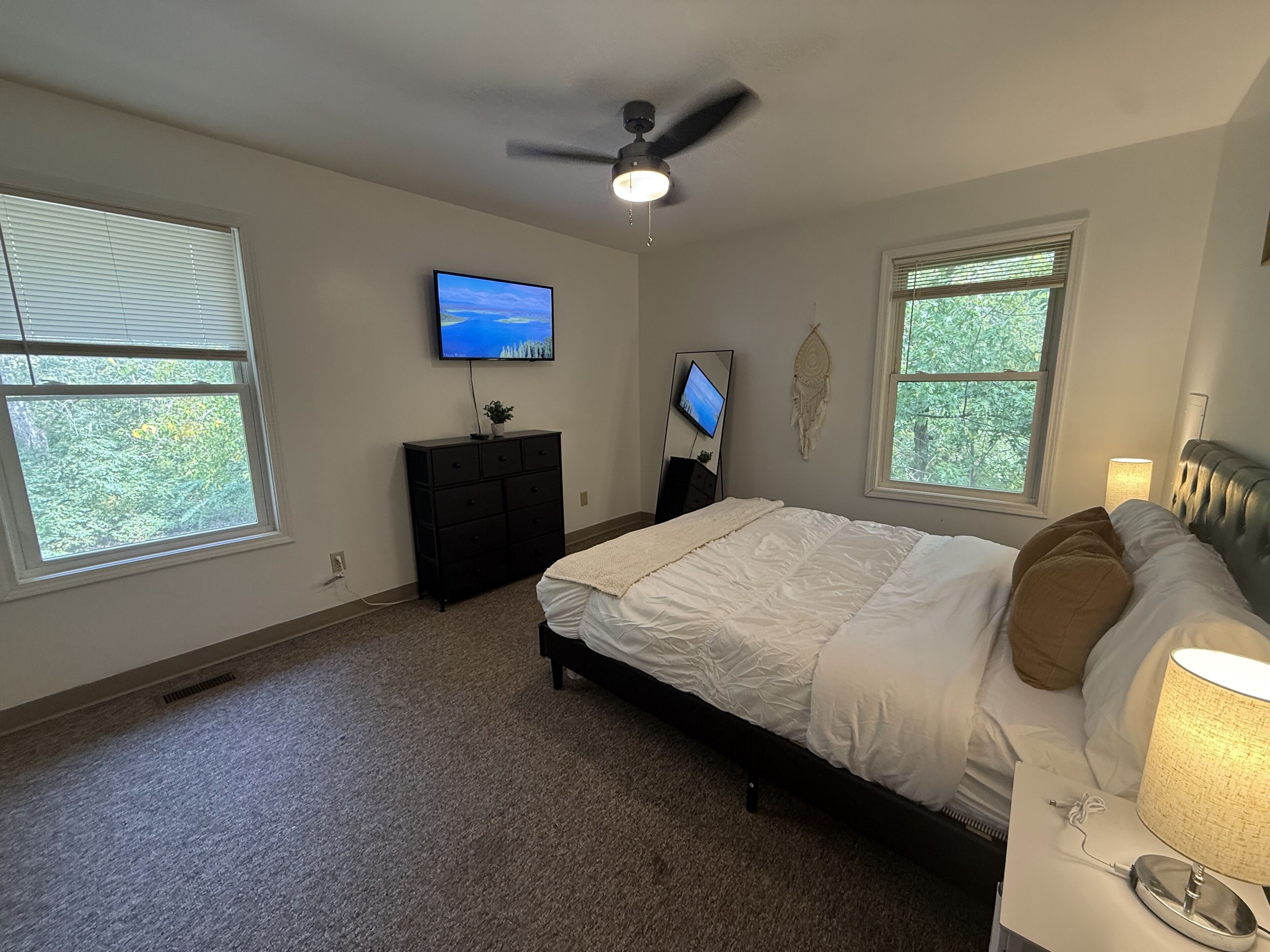 Une chambre à coucher comprenant un lit, un ventilateur de plafond, une télévision, une commode et une fenêtre donnant sur des arbres.
