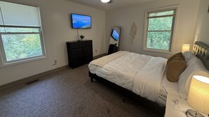 Ein Schlafzimmer mit einem Bett, einem Deckenventilator, einem Fernseher, einer Kommode und einem Fenster mit Blick auf Bäume.
