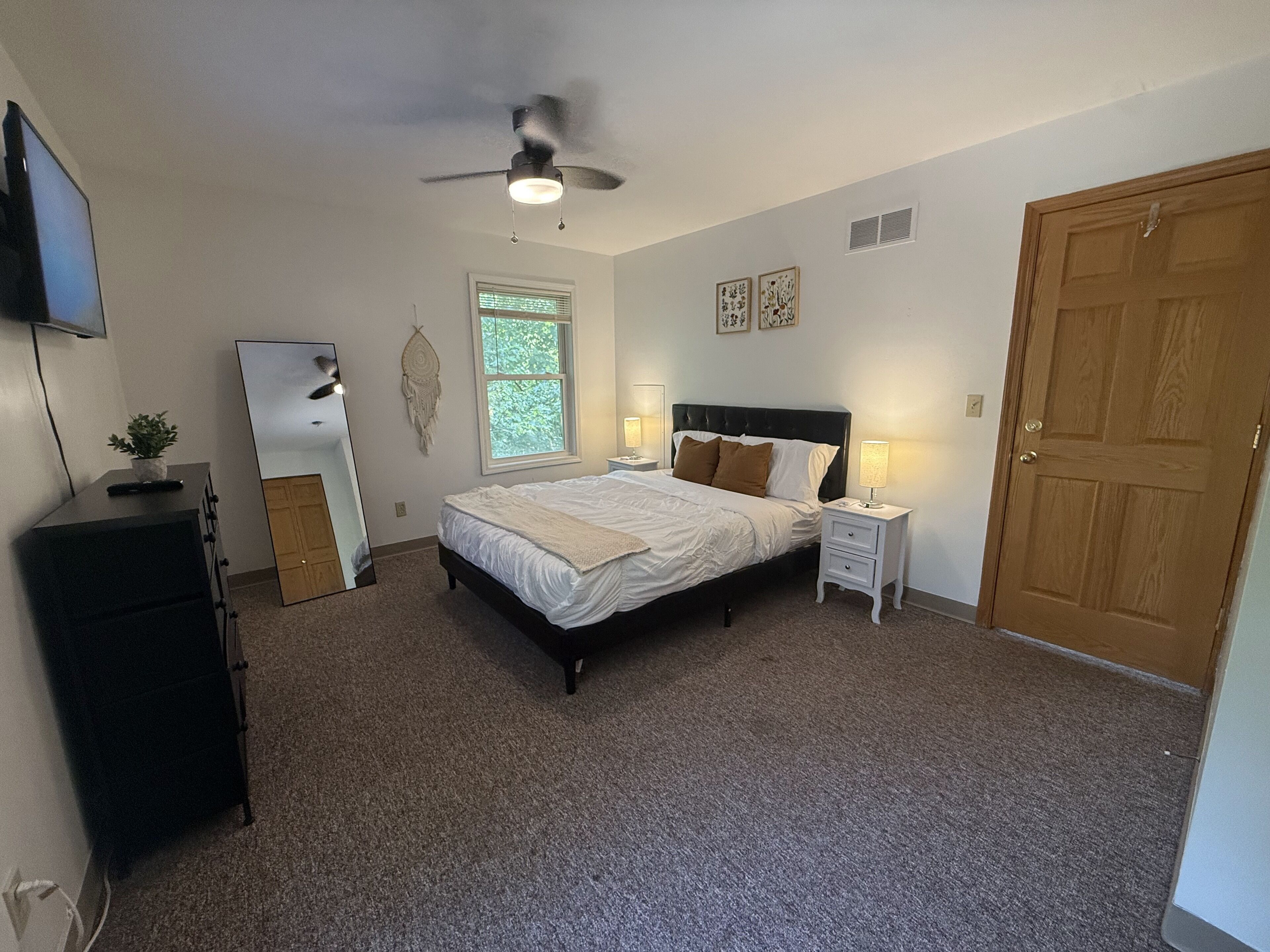Ein Schlafzimmer mit Bett, Nachttischen, Deckenventilator, Fernseher, Kommode, Fenster und Tür.