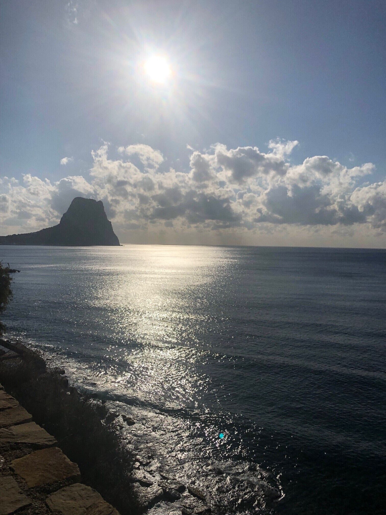 Un paysage côtier avec un ciel dégagé, une grande montagne à l’horizon et une mer calme qui reflète la lumière du soleil.