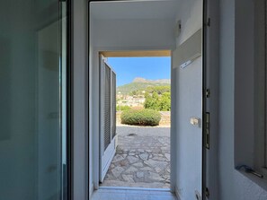Un umbral abierto que da a un patio con suelo de piedra, un arbusto pequeño y vistas de edificios y colinas a lo lejos.
