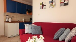 Un salon avec un canapé rouge, une petite table sur laquelle est posé un vase de fleurs, et un coin cuisine avec des meubles bleus et un réfrigérateur blanc.