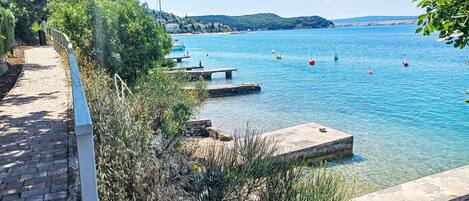 A clear waterfront path with a stone railing, leading to a calm body of water with boats and buoys.