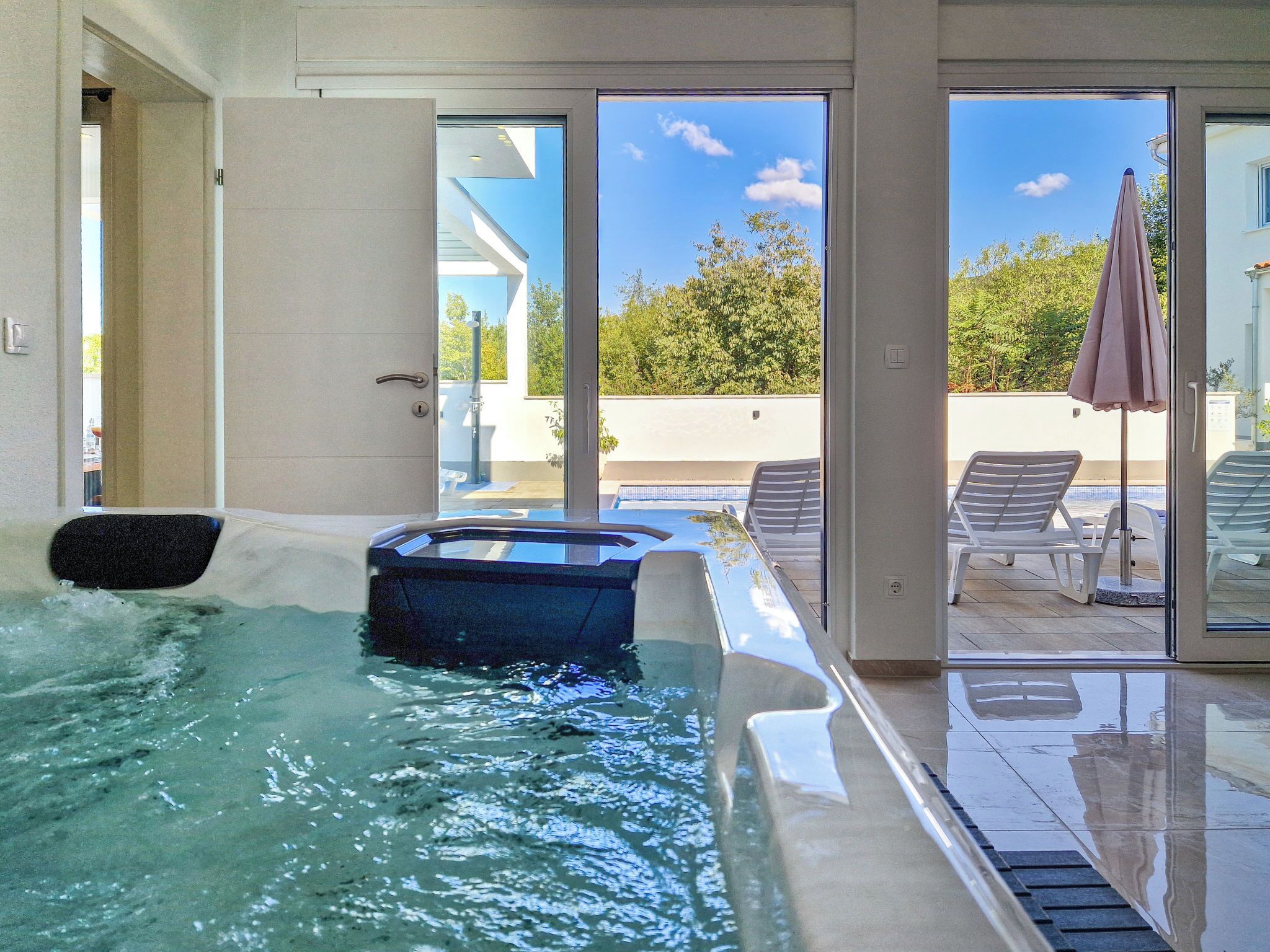 A modern indoor swimming pool with clear glass walls, a lounge area with chairs, and a view of the outdoors.