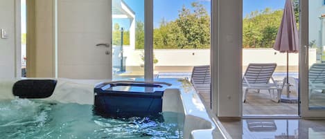 A modern indoor swimming pool with clear glass walls, a lounge area with chairs, and a view of the outdoors.