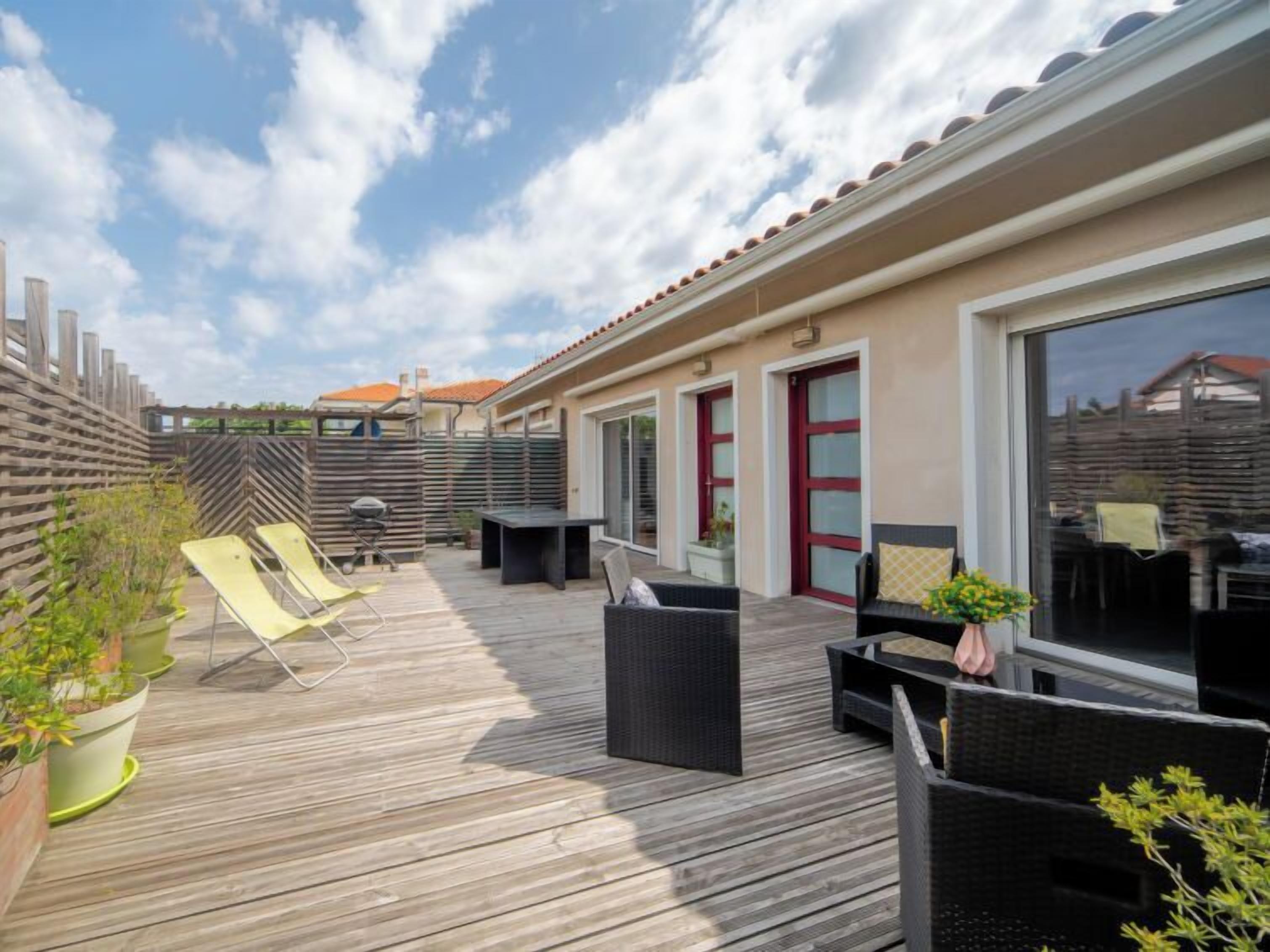 A wooden deck with outdoor furniture, including chairs and a table, and a building with large windows and a red door.