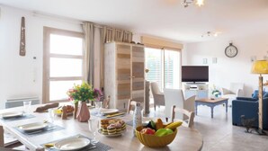 Un comedor con una mesa preparada para dos personas, un frutero y un jarrón con flores. Se observa un televisor al fondo y un reloj en la pared.