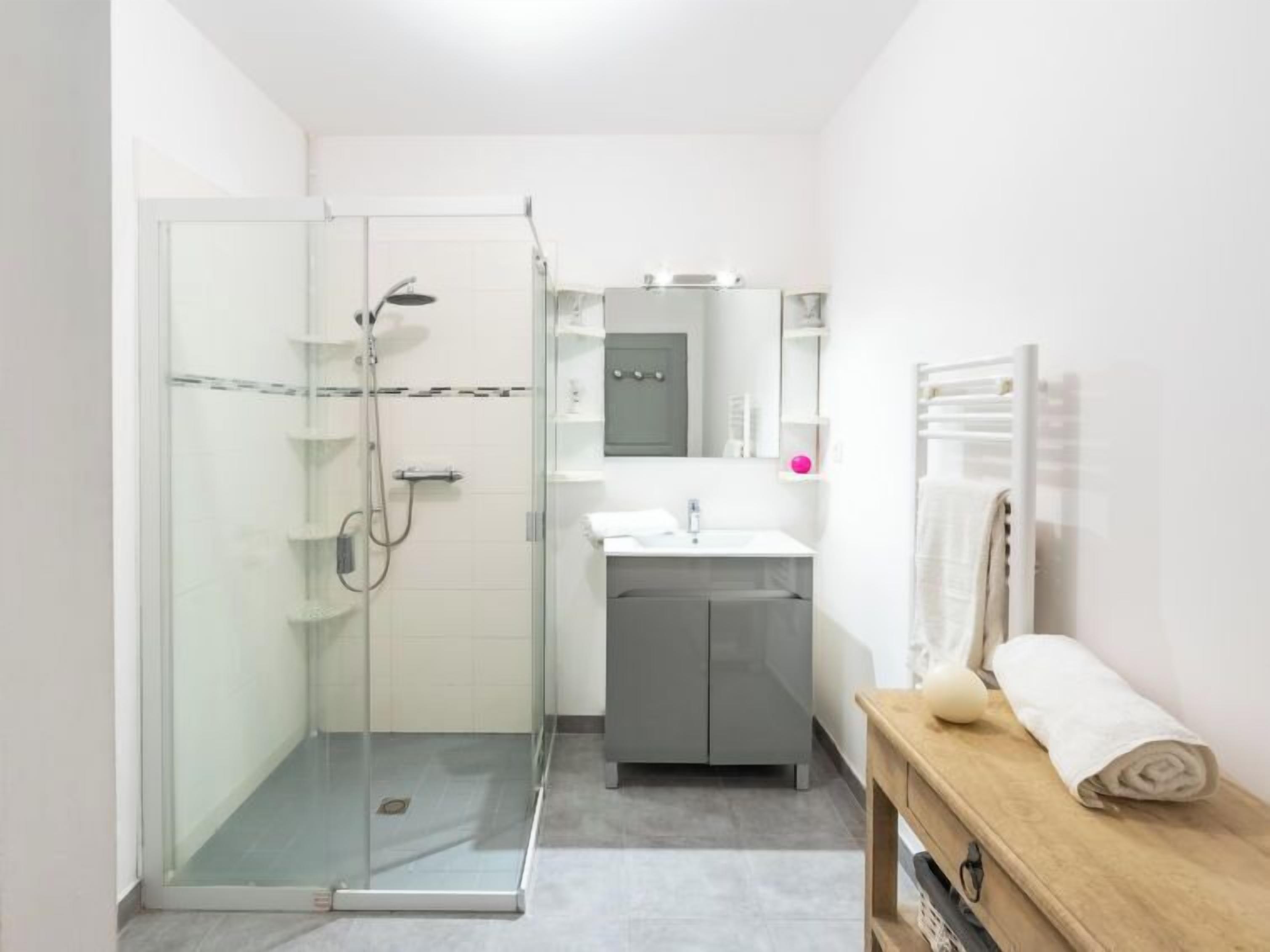 A modern bathroom with a glass shower enclosure, a sink with a mirror, and a wooden bench.