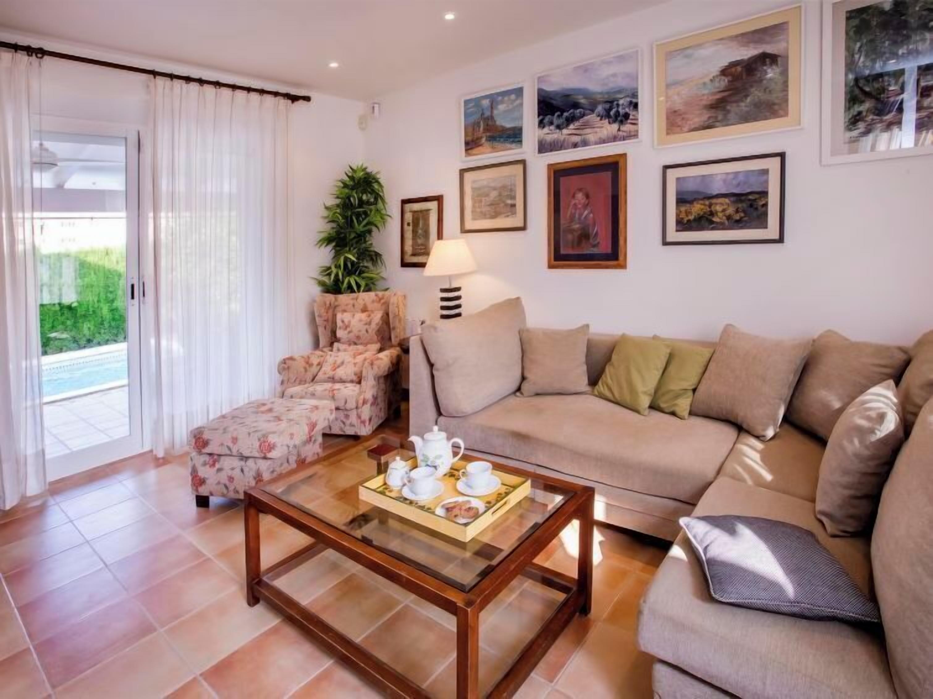 A living room with a sofa, armchair, and coffee table. There are framed pictures on the wall and a plant in the corner.