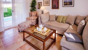 Ein Wohnzimmer mit Sofa, Sesseln und Couchtisch. An den Wänden hängen Bilderrahmen und in der Ecke steht eine Pflanze.