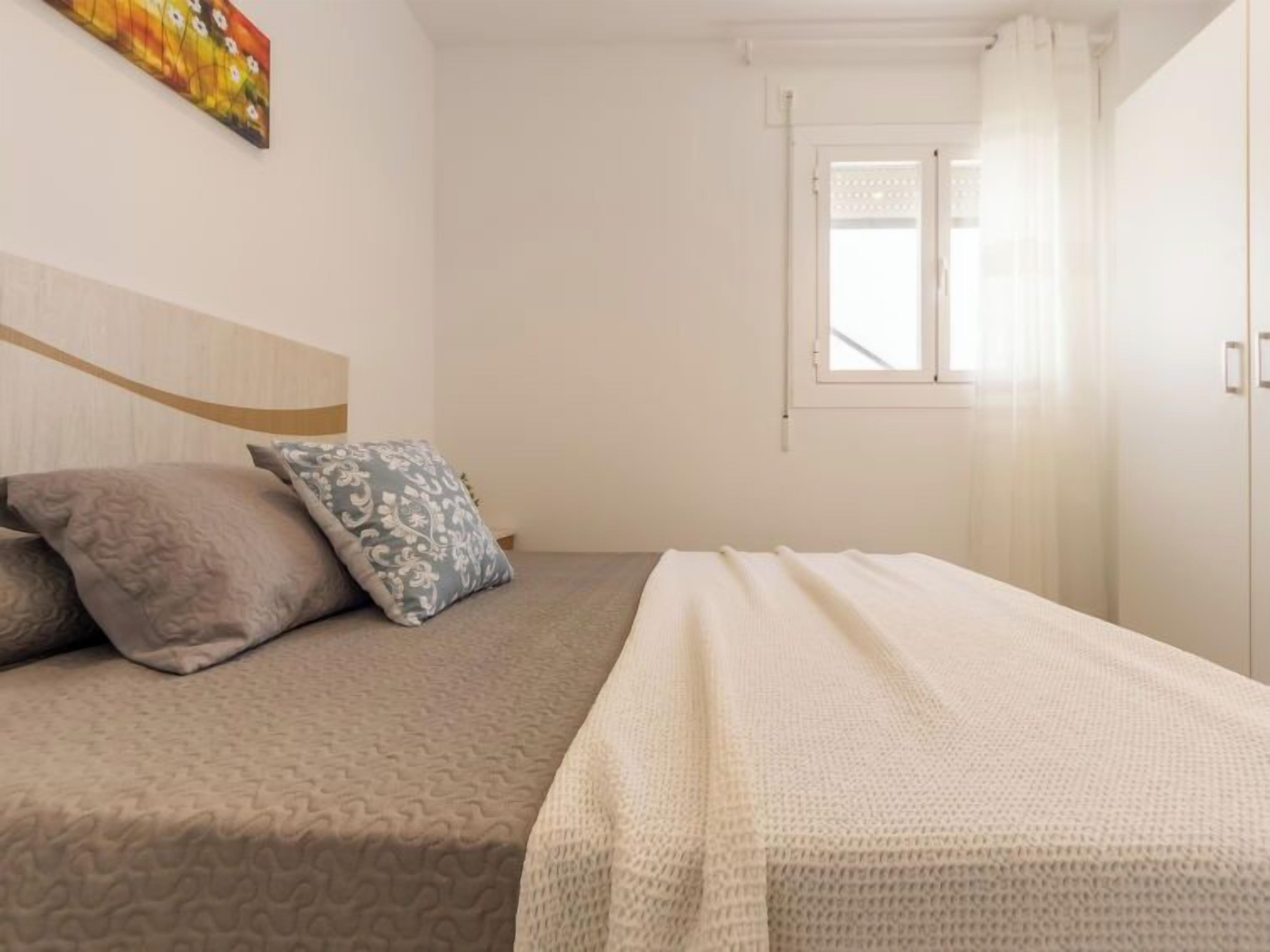 Een netjes opgemaakt bed met een bedspreide en kussens, een raam met gordijnen, en een effen muur met een enkel kunstwerk.