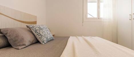 Een netjes opgemaakt bed met een bedspreide en kussens, een raam met gordijnen, en een effen muur met een enkel kunstwerk.