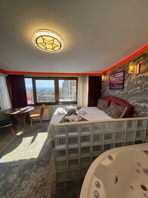 A hotel room with a bed, a desk, a chair, a large window, a jacuzzi, and a marble wall. - Gur Royal Suit Aparts (Istanbul)