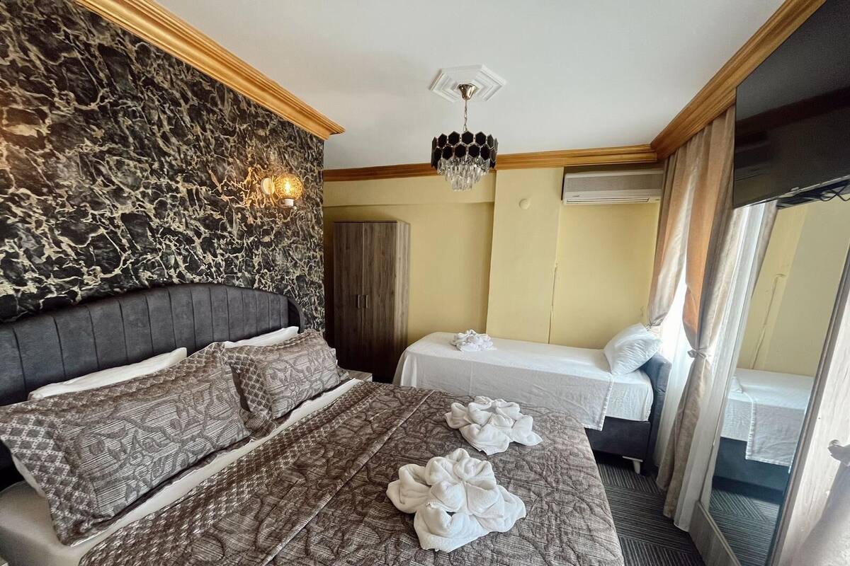 A hotel room with a marble-patterned headboard, a chandelier, a wardrobe, two beds with towels, and a view through a window with curtains.