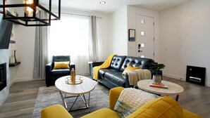 Een moderne woonkamer met een leren zwarte bank, gele kussens, een ronde salontafel en een open haard.