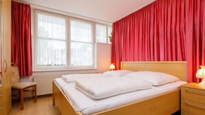 Un dormitorio con una cama doble, dos mesitas de noche con lámparas, una silla y una ventana con cortinas rojas.