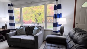A living room with a grey sofa, a black leather couch, and a coffee table. There are two windows with blue and white curtains, and a door with a glass panel. - Hot tub, Nature, Parks and Beaches! Right in between Holland and Grand Haven. (West Olive)