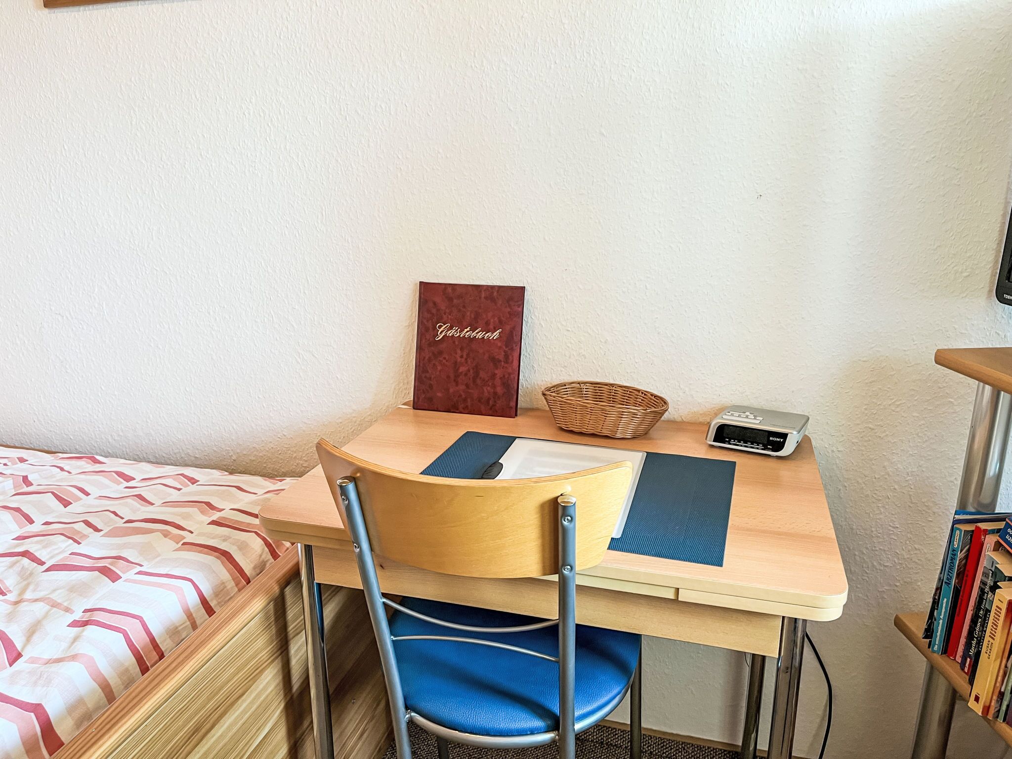 Une petite pièce comprenant un lit, un bureau avec une chaise, un panier, une radio Sony et une bibliothèque.