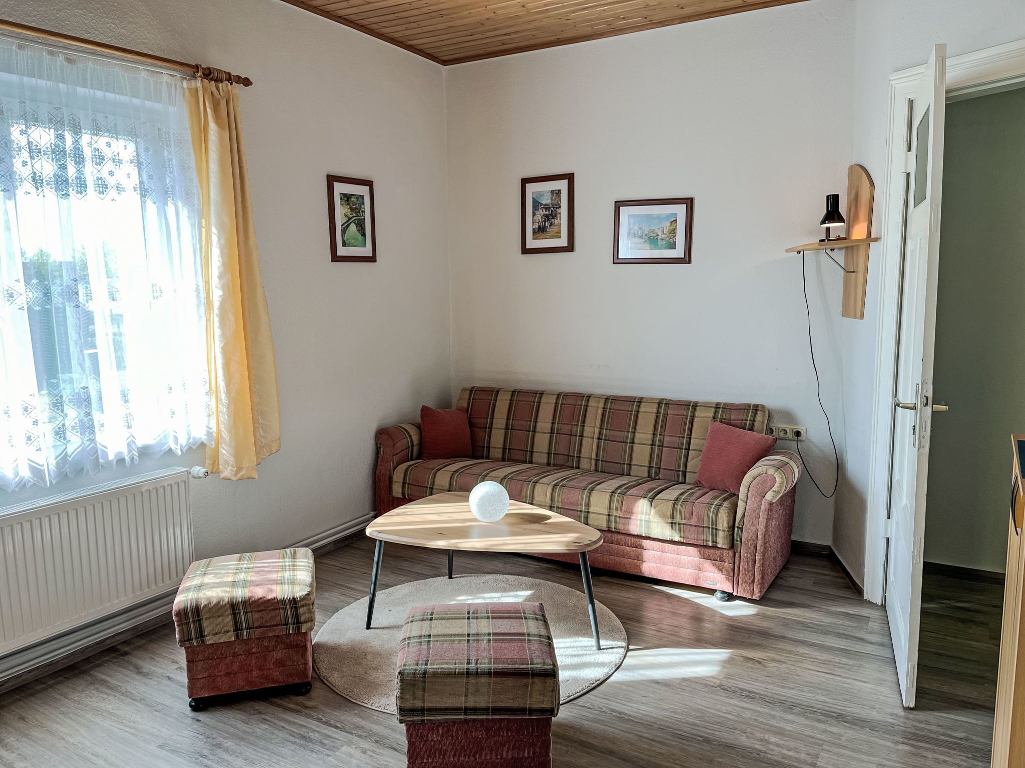 A cozy living room with a plaid sofa, two matching ottomans, a round coffee table, and a window with sheer curtains.