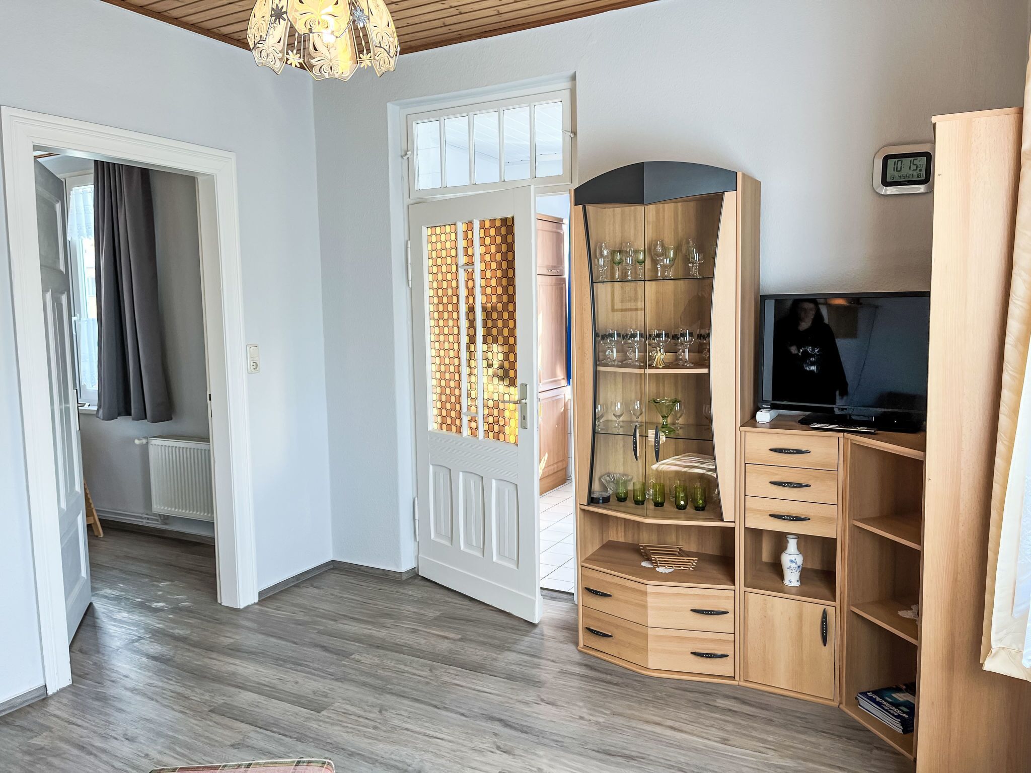 A room with a wooden floor, a white door with a patterned glass panel, a wooden cabinet with glassware, a television, and a clock on the wall.