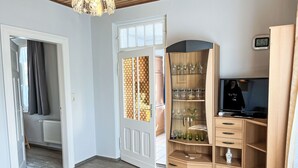 Een kamer met een houten vloer, een witte deur met een ruitpaneel met patroon, een houten kast met glazen, een televisie en een klok aan de muur.