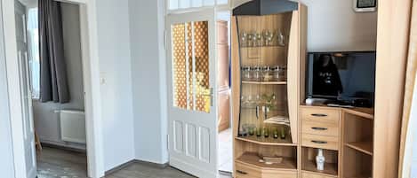 A room with a wooden floor, a white door with a patterned glass panel, a wooden cabinet with glassware, a television, and a clock on the wall.
