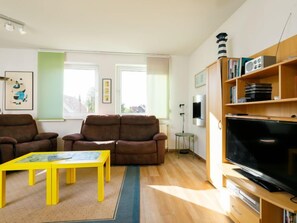 Un salon avec deux canapés marron, une table basse jaune, un téléviseur posé sur un meuble en bois et une bibliothèque contenant divers objets.