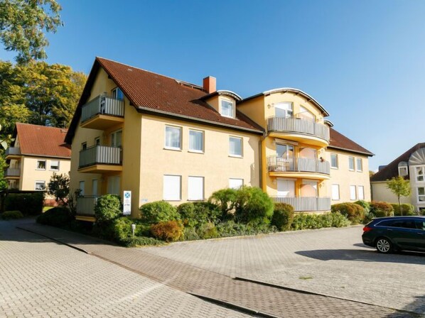 Un edificio residencial de varios pisos con balcones, entrada pavimentada y zonas verdes circundantes.