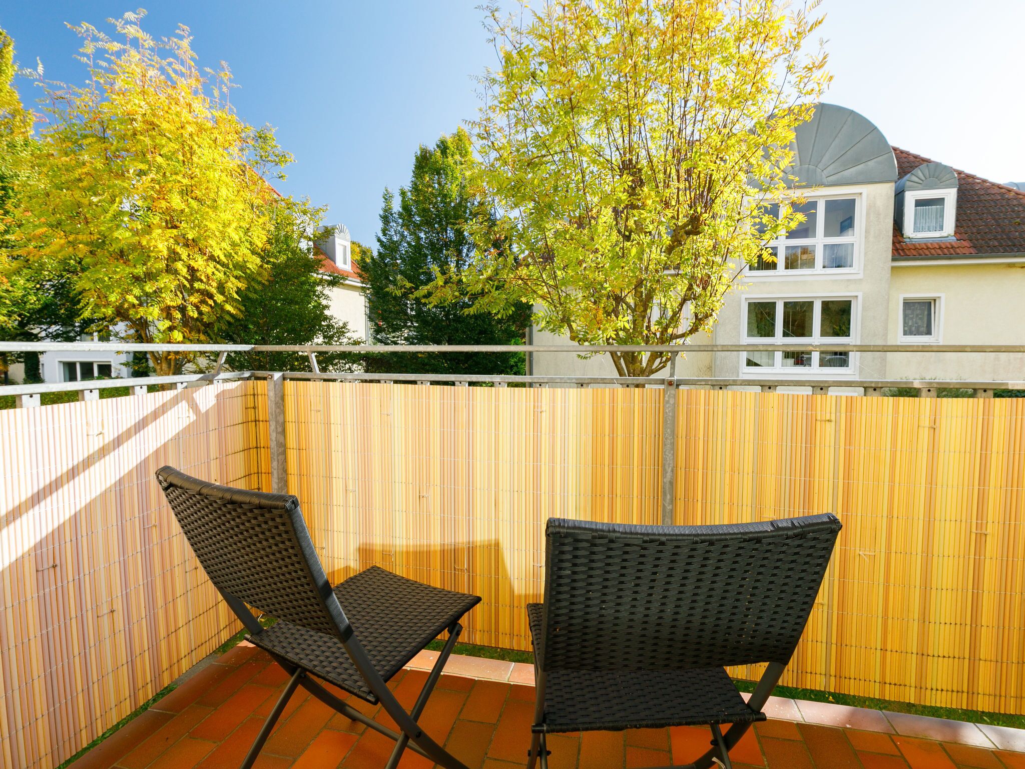 Un balcon avec deux fauteuils en osier, une clôture en bambou et une vue sur un immeuble avec un toit rouge.