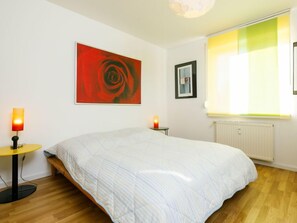 Une chambre avec un grand lit, un tableau abstrait rouge, un tableau encadré, une table avec une lampe et une fenêtre avec des stores.