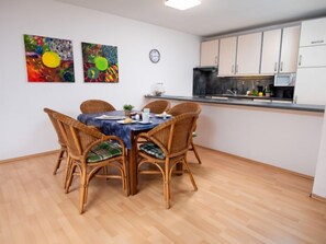 Een eethoek met een tafel voor vier personen, omringd door rieten stoelen met groene kussens. Twee abstracte schilderijen hangen aan de muur, een klok, en een keuken met witte kasten en een zwart aanrecht.