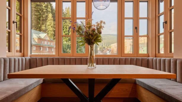 A dining area with a wooden table, cushioned benches, and large windows offering a view of buildings and trees. - Summit View Lounge & Alcove by Interhome (Pec pod Snezkou)
