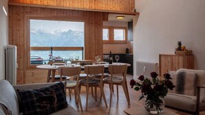 A cozy room with a wooden ceiling, a dining table with chairs, a sofa, and a view of mountains through a large window. - Haut de Cry 16 by Interhome (Nendaz)