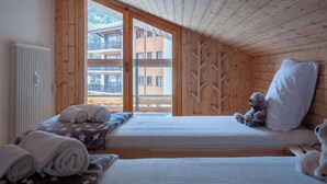 A cozy room with wooden walls and a sloped ceiling, featuring two beds with pillows and a teddy bear, a radiator, and a view of a building through a window. - Haut de Cry 16 by Interhome (Nendaz)