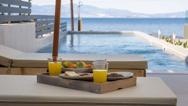 Bandeja de desayuno junto a la piscina con frutas y zumo, una tumbona y vistas al mar y las montañas.