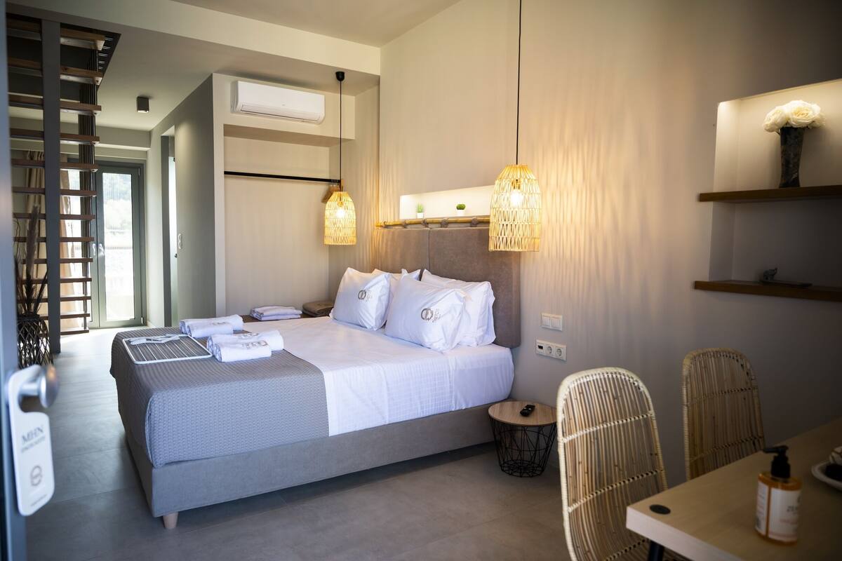 Una habitación de hotel moderna con una cama grande, una mesa de comedor, sillas de mimbre y una mesita auxiliar.