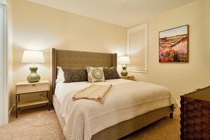 A neatly arranged bedroom with a large bed, two bedside tables with lamps, a decorative pillow, a wall-mounted painting, and a small wooden cabinet. - The Mariposa-Valerian Bundle at Academy Place (Crested Butte)