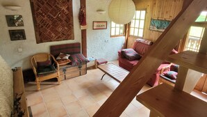 Interior em madeira com teto alto, soalho em azulejos e escadaria. Há duas cadeiras, uma mesa pequena e uma estante fixada na parede.