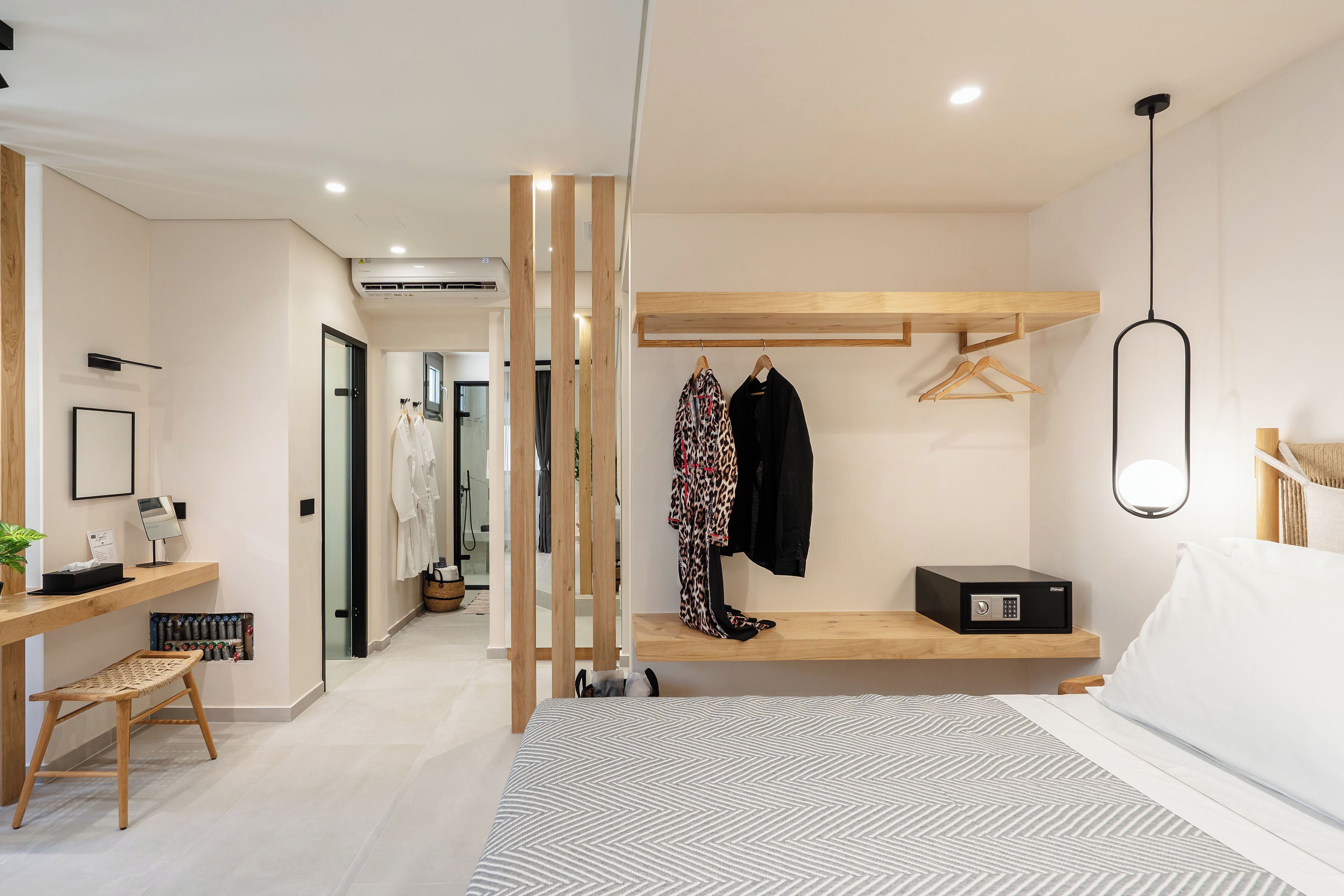 Une chambre d’hôtel moderne avec un lit, une table de chevet et un banc en bois. On y trouve un porte-manteau avec des vêtements et une petite boîte noire posée sur une étagère.