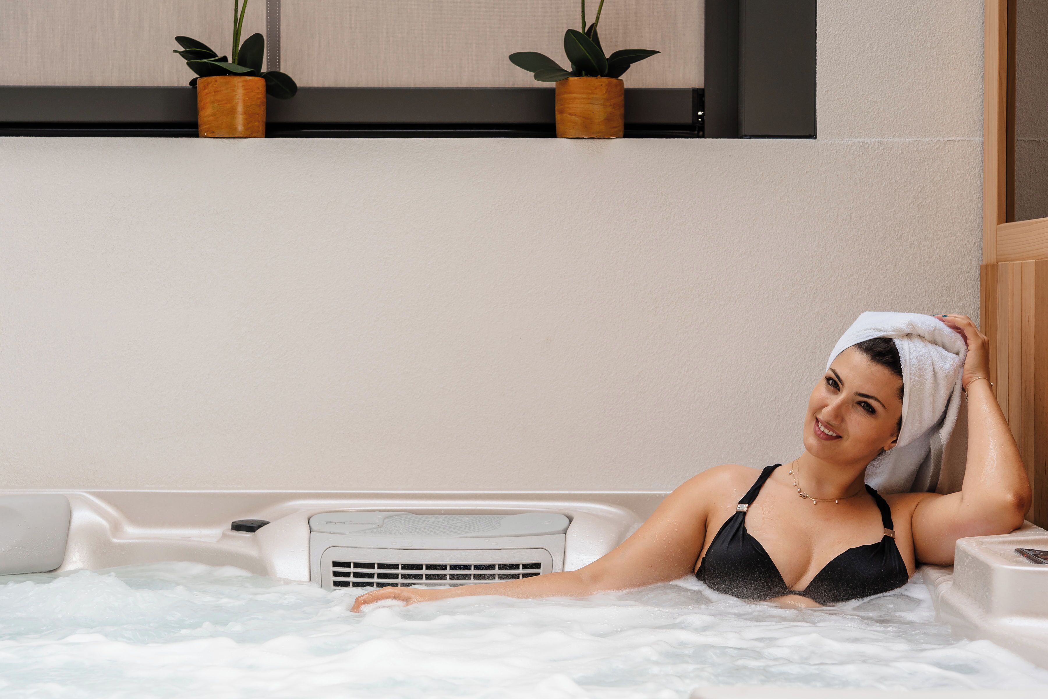 Eine Person in einer Badewanne, ein Handtuch auf dem Kopf, eine weiße Wand und zwei Blumentöpfe auf einem Regal.