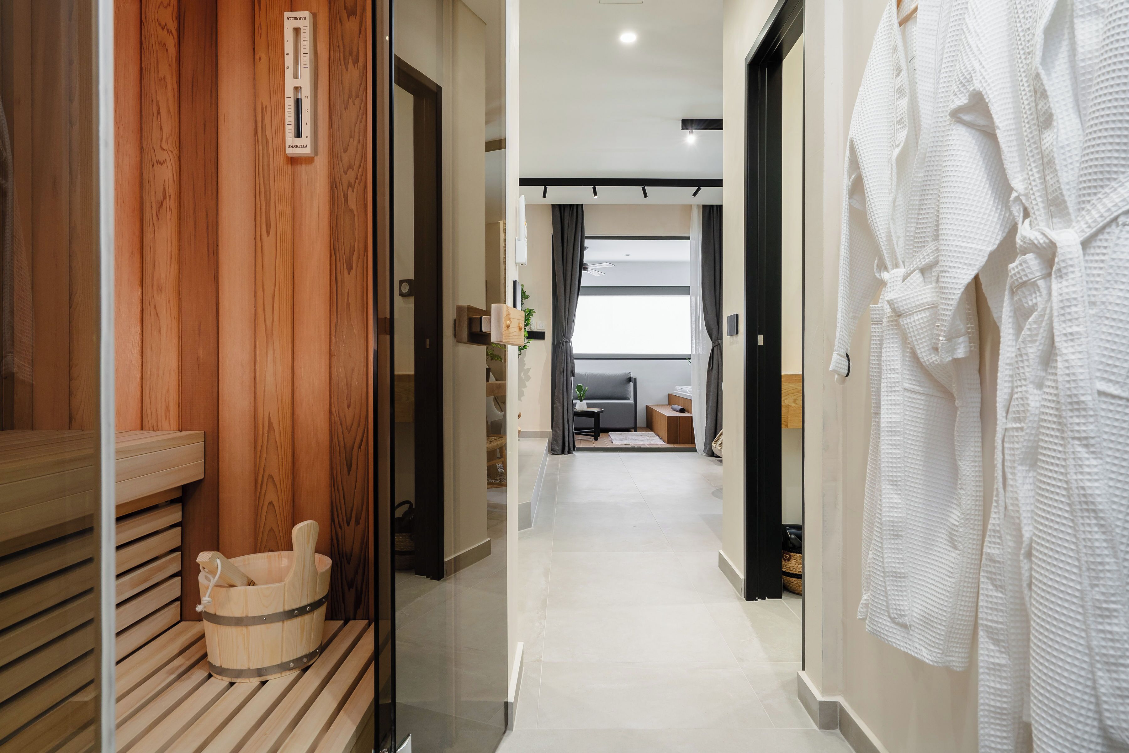 A hallway with a sauna, a wooden bench, a white robe rack, and a glimpse of a living area with a sofa and a plant.