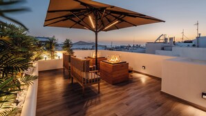 A rooftop terrace with a wooden deck, a large umbrella, a fire pit, and a view of the cityscape. - Legato Spa Suites (Naxos)