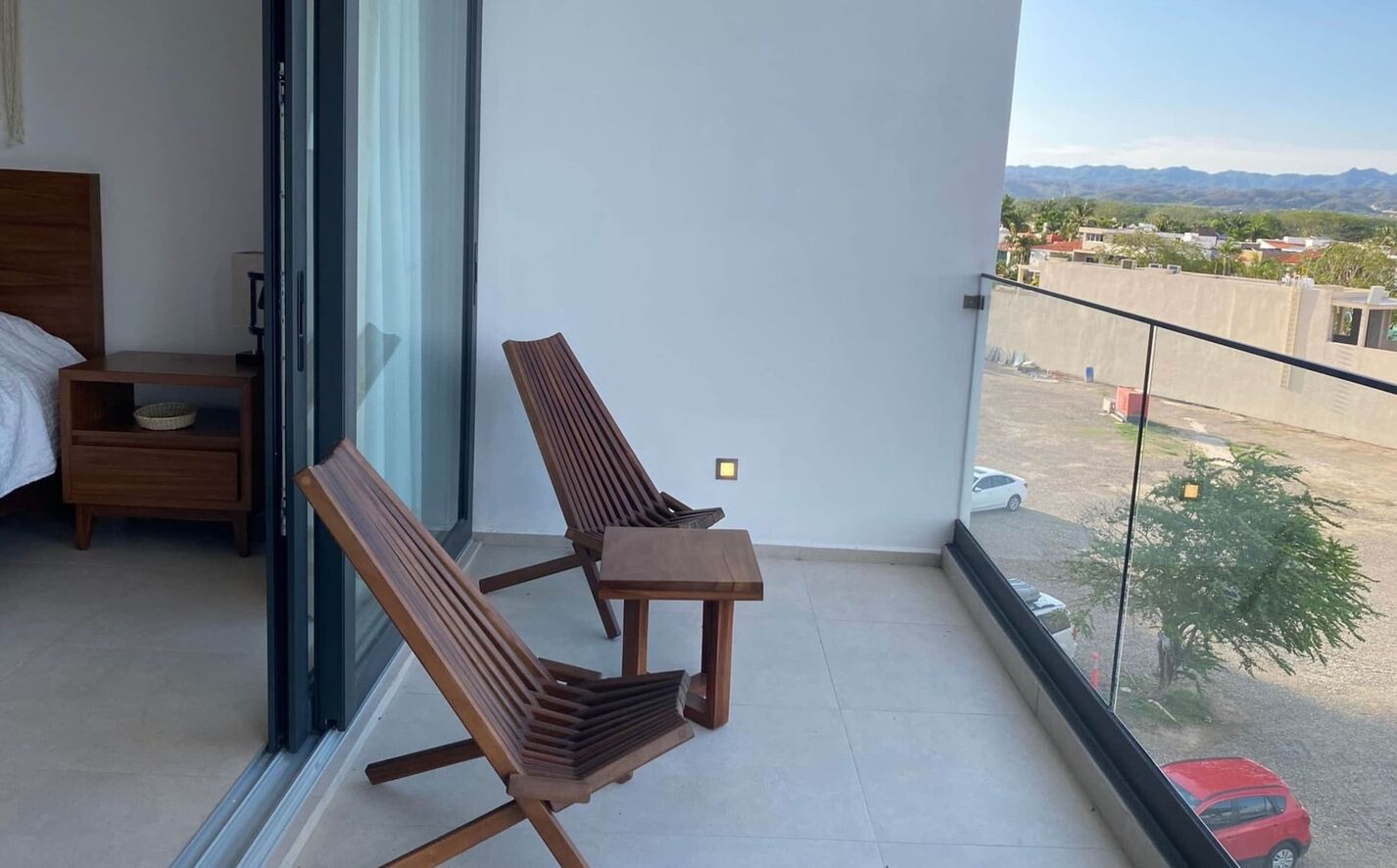 Un balcon avec deux chaises en bois et une petite table, une table de chevet en bois et un lit recouvert d’une courtepointe blanche.