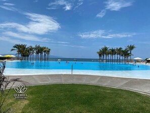 Una amplia piscina al aire libre con palmeras y sombrillas, rodeada de césped y un cielo azul despejado.
