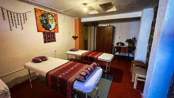 Two beds are covered with patterned blankets, accompanied by a painting and a small table with a chair.