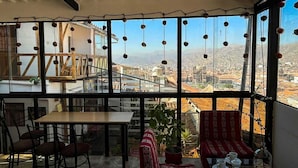 A dining area with a table and chairs, a red patterned sofa, and a view of a cityscape through large windows. - Casa Cusco guest house (Cusco)