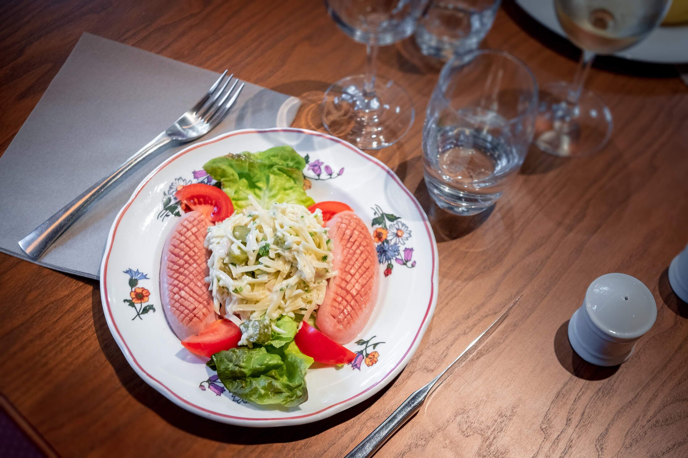 Ein Teller mit Essen, einer Gabel, einem Messer und leeren Weingläsern auf einem Holztisch.